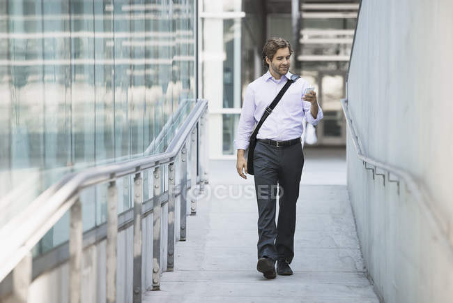 Giovane uomo che trasporta borsa per computer con cinturino e utilizzando smartphone sulla passerella della città . — Foto stock