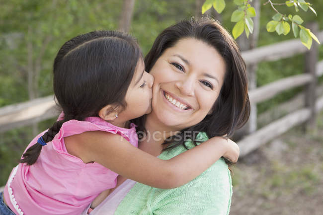 Mutter trägt Tochter, lächelt und bekommt Kuss von Mädchen im Park. — Stockfoto