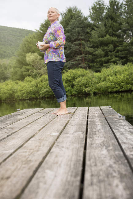 Low angle view of woman standing on lake dock and holding coffee cup. — Stock Photo