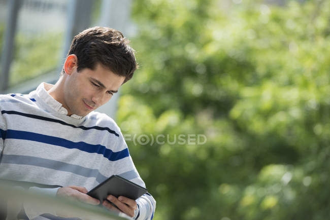 Молодий чоловік спирається на перила в парку і використовує цифровий планшет . — стокове фото