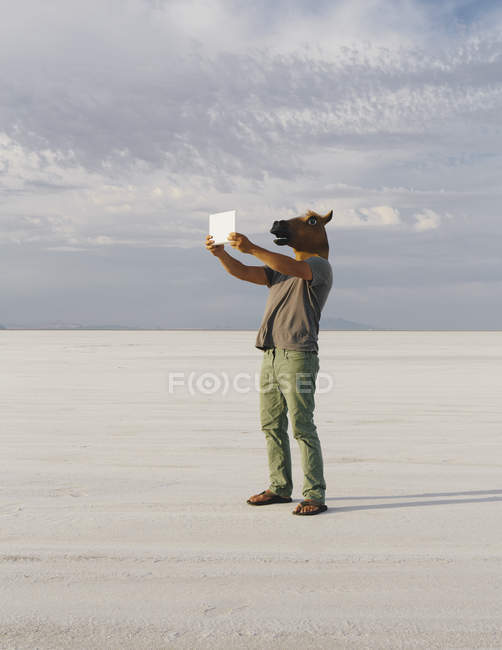 Hombre con máscara de caballo tomando fotos con dispositivo de tableta en Bonneville Salt Flats, Utah, EE.UU. . - foto de stock
