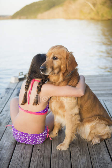 Pre-adolescent girl in swimwear hugging golden retriever dog while sitting on a jetty. — Stock Photo