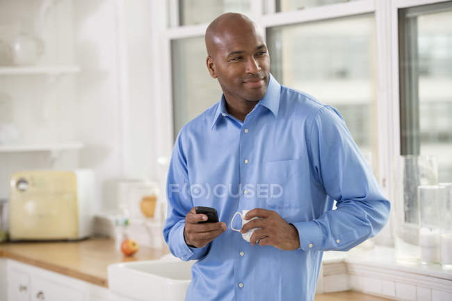 Mann im blauen Hemd hält Smartphone und Tasse Kaffee in Büroküche. — Stockfoto