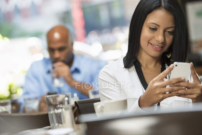 Woman smiling while using smartphone at coffee shop table with man drinking in background. — Stock Photo