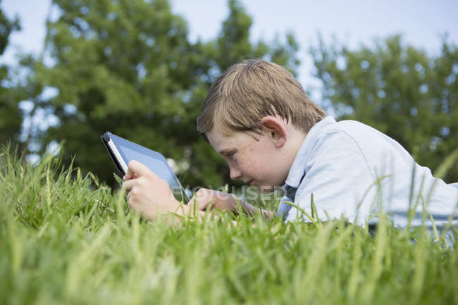 Хлопчик початкового віку лежить на траві і використовує цифровий планшет . — стокове фото