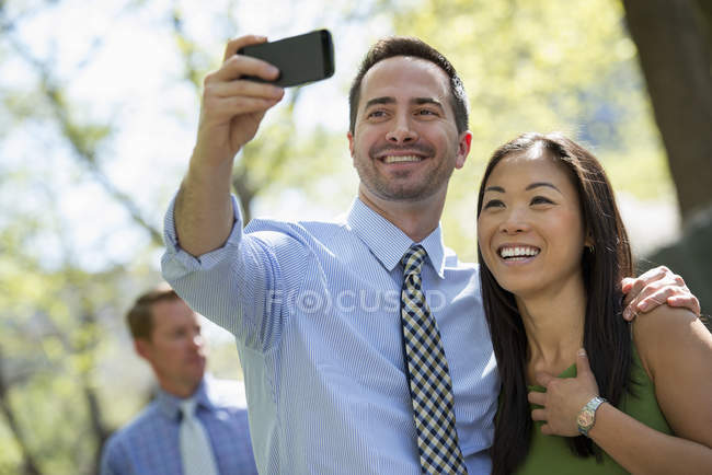 Casal tirando selfie com smartphone com homem de pé no fundo . — Fotografia de Stock