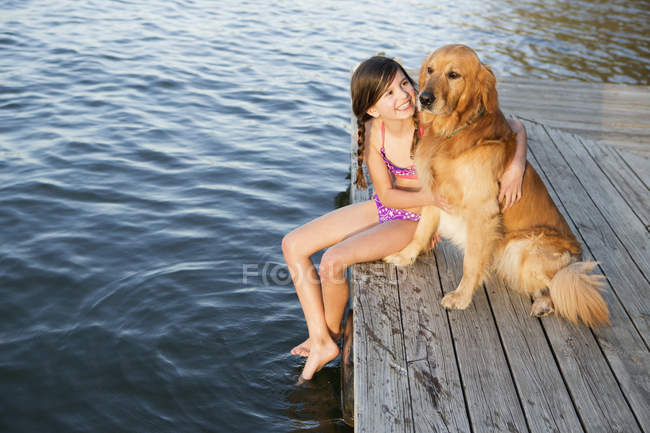 Pre-adolescent girl in swimwear with golden retriever dog sitting on jetty by lake. — Stock Photo