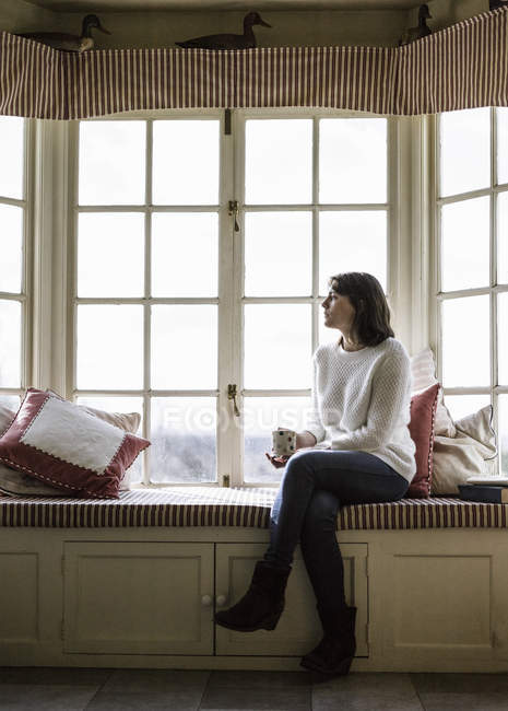 Mulher pensativa sentada na soleira da janela com xícara de chá . — Fotografia de Stock
