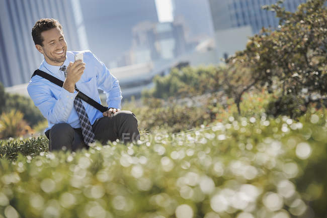 Молодой человек сидит на скамейке в парке, улыбается и пользуется мобильным телефоном . — стоковое фото