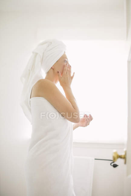 Mujer envuelta en toalla blanca de pie en el baño y aplicando crema . - foto de stock