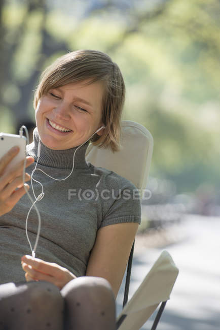 Femme assise dans une chaise de camping dans un parc et écoutant de la musique dans des écouteurs . — Photo de stock