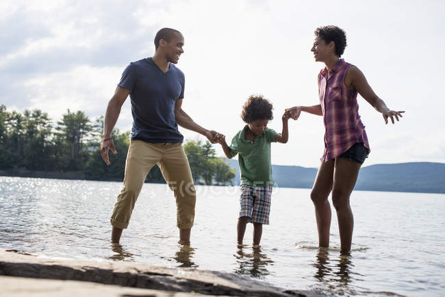 Padres e hijo tomados de la mano y caminando en el agua del lago en verano . - foto de stock