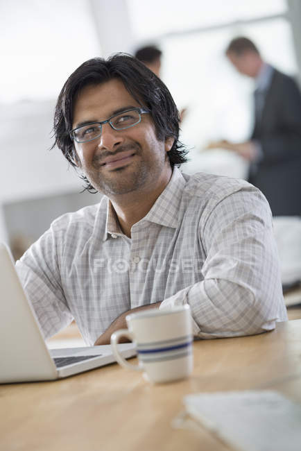 Hombre en la oficina usando el ordenador portátil en la mesa con una taza de café y colegas hablando en segundo plano . - foto de stock