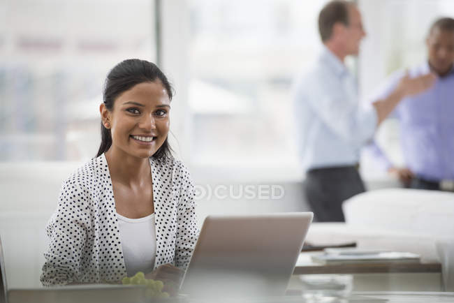 Giovane donna sorridente mentre si siede alla scrivania con computer portatile con gli uomini che parlano in background . — Foto stock