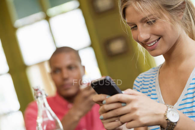 Frau checkt Smartphone mit Mann im Hintergrund in Café. — Stockfoto