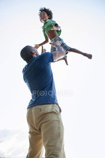 Low angle view of father playing and lifting elementary age boy outdoors. — Stock Photo