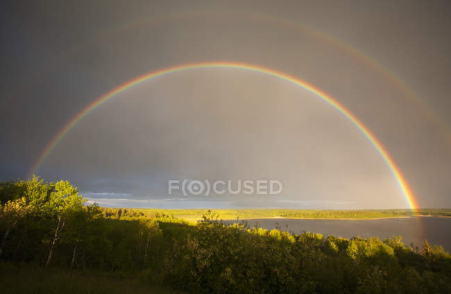 Double rainbow in sky arching over forest and lake in Canada. — Stock Photo