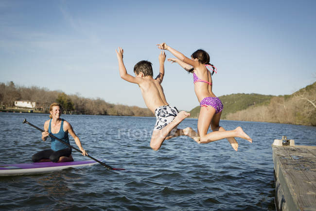 Les enfants sautent dans l'eau de la jetée avec une femme sur la planche à pagaie regarder . — Photo de stock