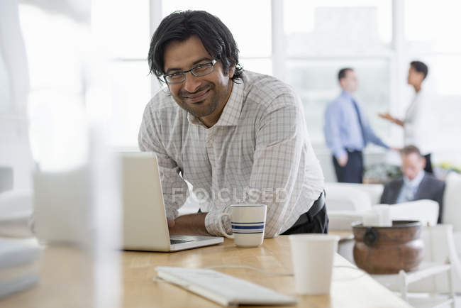 Älterer Mann lehnt mit Laptop am Schreibtisch im Büro, während Kollegen im Hintergrund reden. — Stockfoto