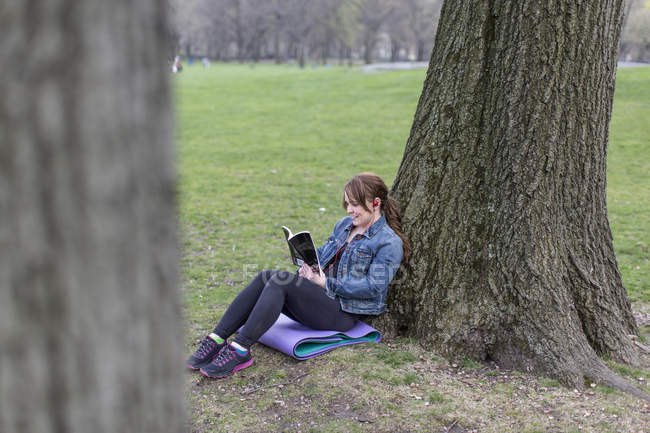 Молодая женщина, опирающаяся на дерево и чтение в Центральном парке, Нью-Йорк, США . — стоковое фото