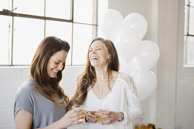 Dos mujeres de pie lado a lado y sosteniendo bebidas en la habitación decorada con globos blancos . - foto de stock