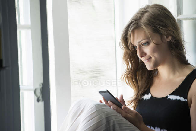 Junge Frau sitzt am Fenster und schaut aufs Smartphone. — Stockfoto