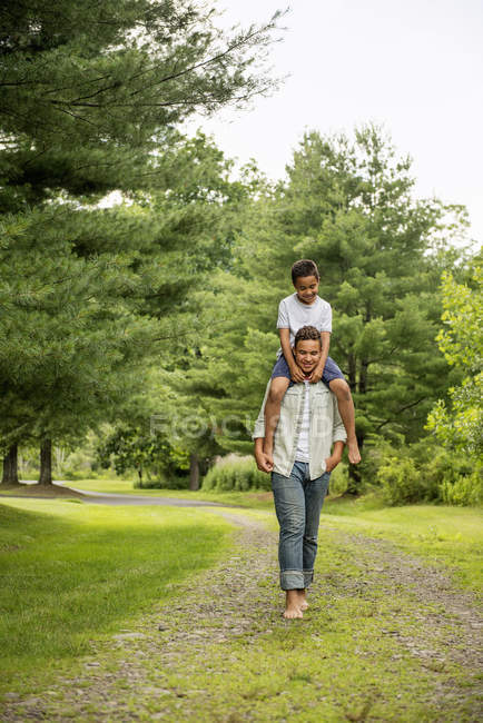 Pre-adolescent boy carrying brother on shoulders while walking on country path. — Stock Photo