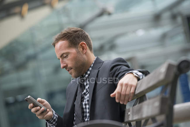 Чоловік у діловому костюмі з коротким волоссям за допомогою смартфона на вуличній лавці . — стокове фото