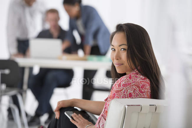 Asian woman relaxing on office sofa with colleagues using laptop in background. — Stock Photo