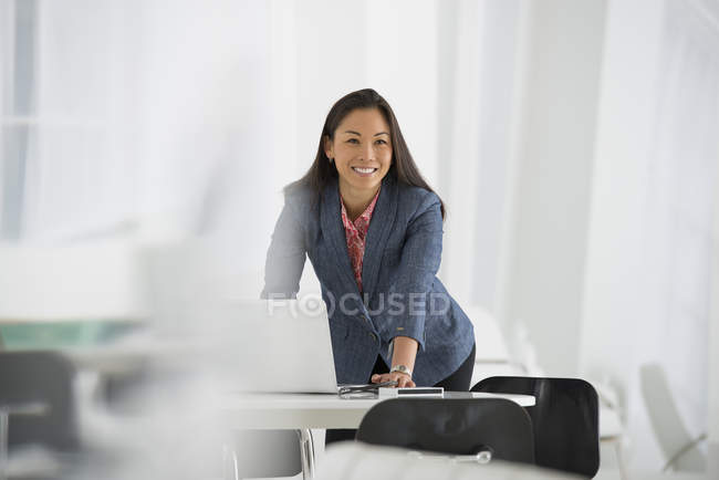 Donna d'affari sorridente appoggiata alla scrivania con laptop in ufficio . — Foto stock