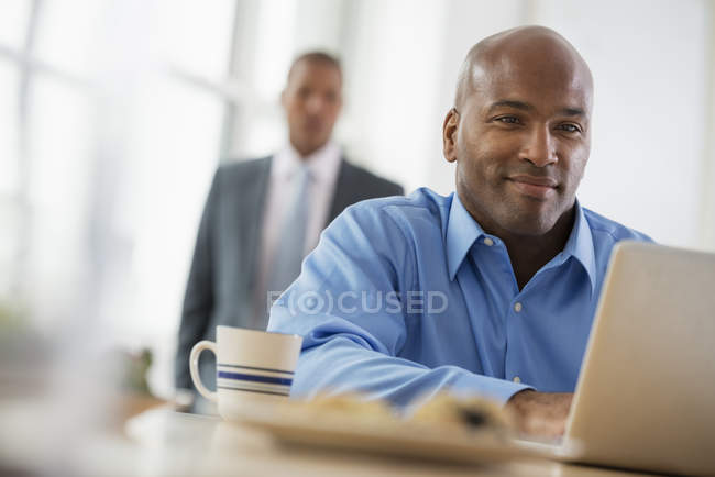 Mann sitzt am Schreibtisch und benutzt Laptop mit Mitarbeiter im Hintergrund. — Stockfoto