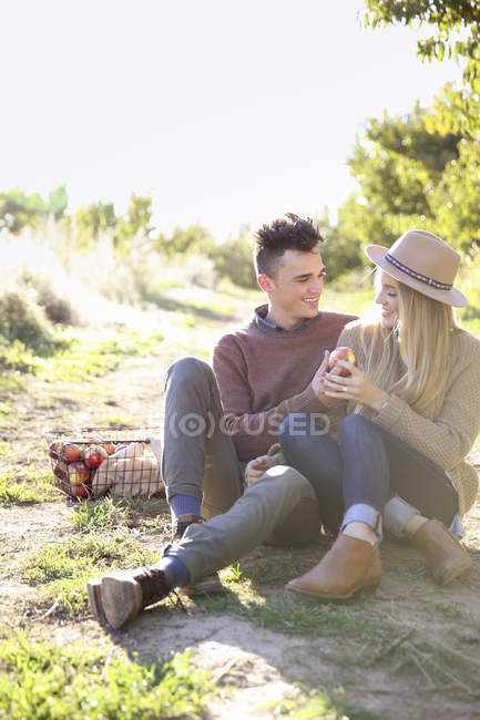 Junges Paar sitzt auf dem Boden mit einem Korb voller Äpfel im Obstgarten. — Stockfoto