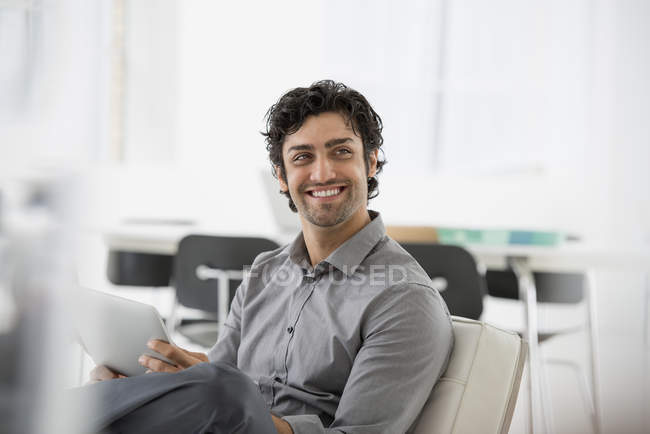 Молодий бізнесмен посміхається і тримає цифровий планшет в офісному кріслі . — стокове фото