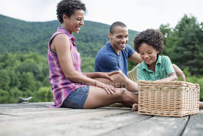 Familie mit Grundschulkind sitzt auf Seebrücke mit Picknickkorb. — Stockfoto