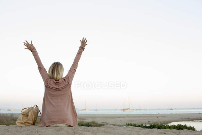 Mulher loira sentada na praia de areia com os braços levantados . — Fotografia de Stock
