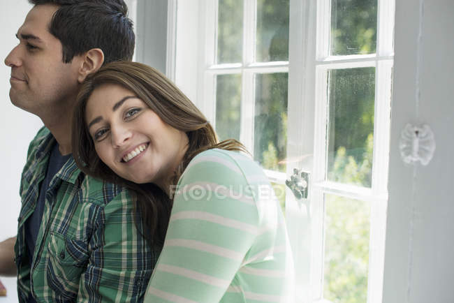 Pareja joven abrazándose en el interior por la ventana, mujer apoyada en el hombro masculino . - foto de stock