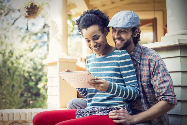 Pareja sentada en escalones de porche, sonriendo y compartiendo tableta digital . - foto de stock