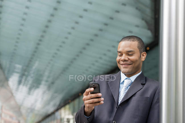Homme d'affaires en costume debout dans la rue, souriant et utilisant un smartphone . — Photo de stock