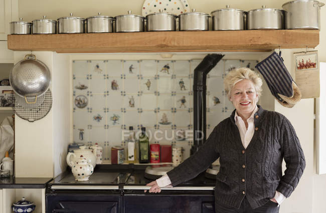 Mature woman standing by range cooker in kitchen interior. — Stock Photo