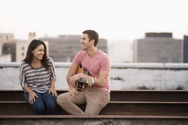Чоловік тримає гітару і сидить зі усміхненою жінкою на терасі на даху з видом на місто в сутінках . — стокове фото