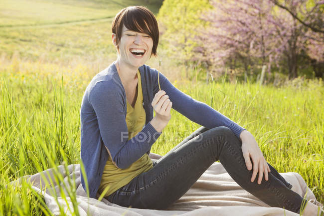 Young woman sitting on grassy field on blanket with dandelion stem. — Stock Photo