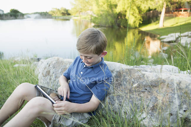Junge im Grundschulalter sitzt am Wasser und nutzt digitales Tablet. — Stockfoto