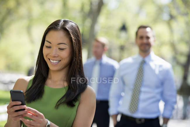 Mid adult woman using smartphone with businessmen in background on city street. — Stock Photo