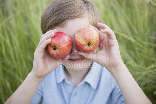 Elementary age boy holding red apples over eyes. — Stock Photo