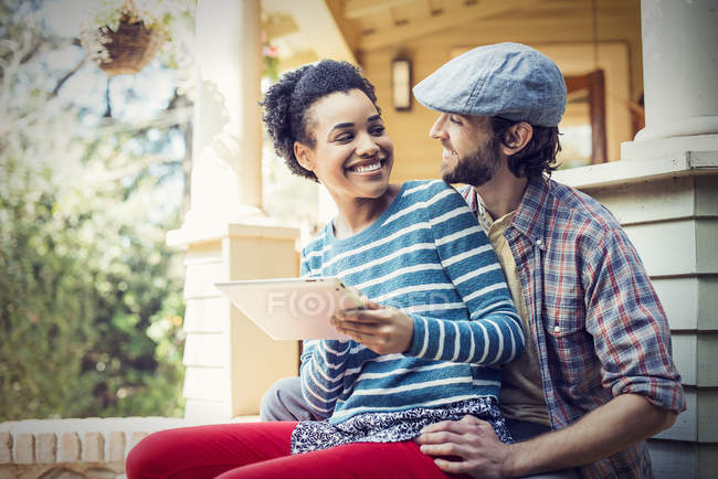 Couple sitting on porch steps with digital tablet, smiling and looking at each other. — Stock Photo