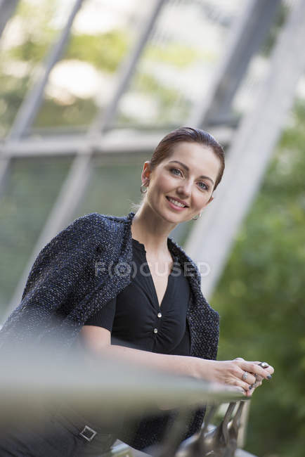 Businesswoman en chaqueta gris con el pelo hacia arriba apoyándose en barandilla en la ciudad . - foto de stock