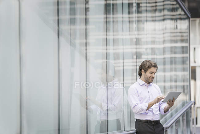 Junger Mann steht vor Gebäude mit gläsernen Außenpaneelen und nutzt digitales Tablet. — Stockfoto