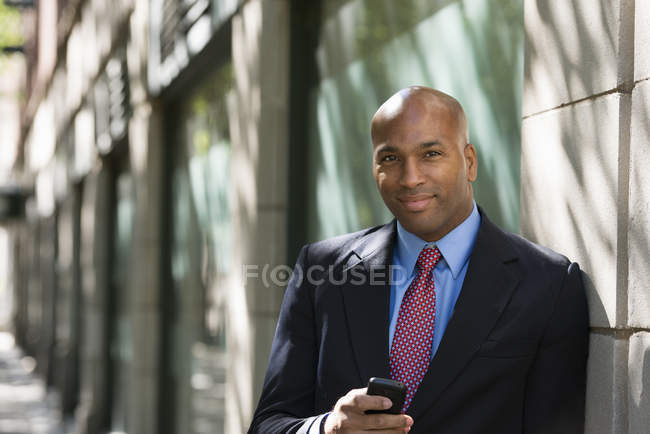 Бизнесмен в костюме и галстуке держит телефон и смотрит в камеру, опираясь на стену на улице . — стоковое фото