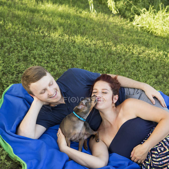 Chihuahua cão lambendo mulher descansando no tapete azul com o homem no parque . — Fotografia de Stock