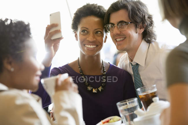 Man and woman posing for selfie at restaurant table with friends. — Stock Photo